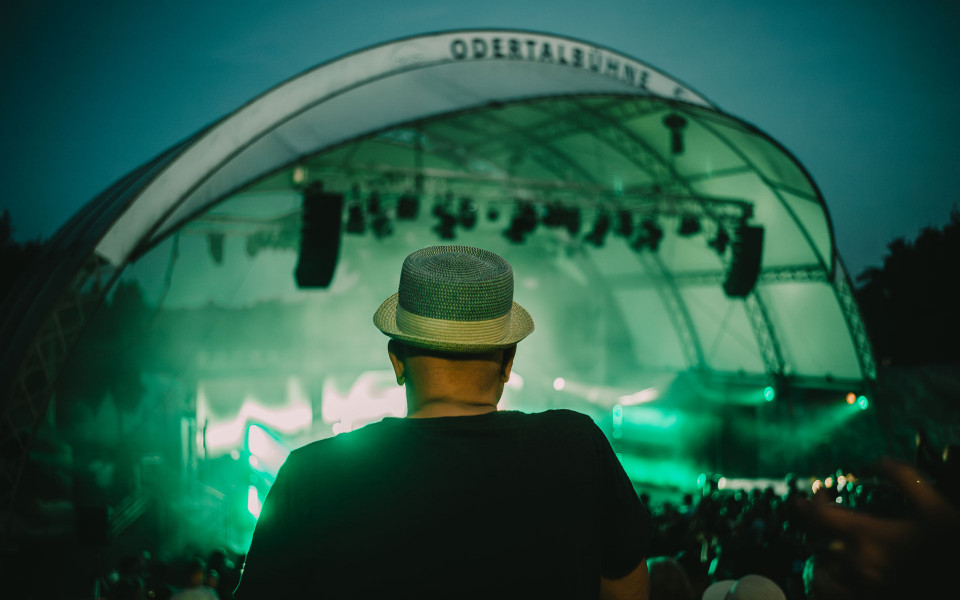 Eine Person mit Strohhut steht von hinten im Publikum vor einer großen, grün beleuchteten Konzertbühne. Nebel, Scheinwerfer und die gewölbte Bühnenkonstruktion erzeugen eine stimmungsvolle Festivalatmosphäre.