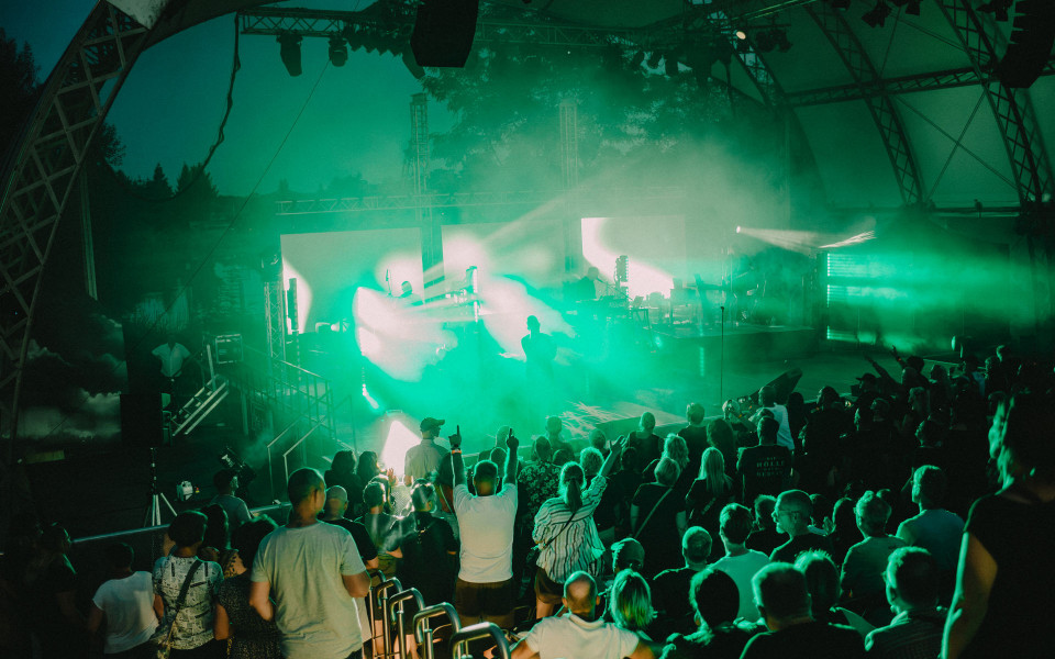 Ein Publikum steht und sitzt in einer Freiluftarena vor einer Konzertbühne, die in intensives grünes Licht und Nebel getaucht ist. Auf der Bühne sind Scheinwerfer, große Bildschirme und Musiker im Dunst zu sehen, während einige Zuschauer die Arme heben.