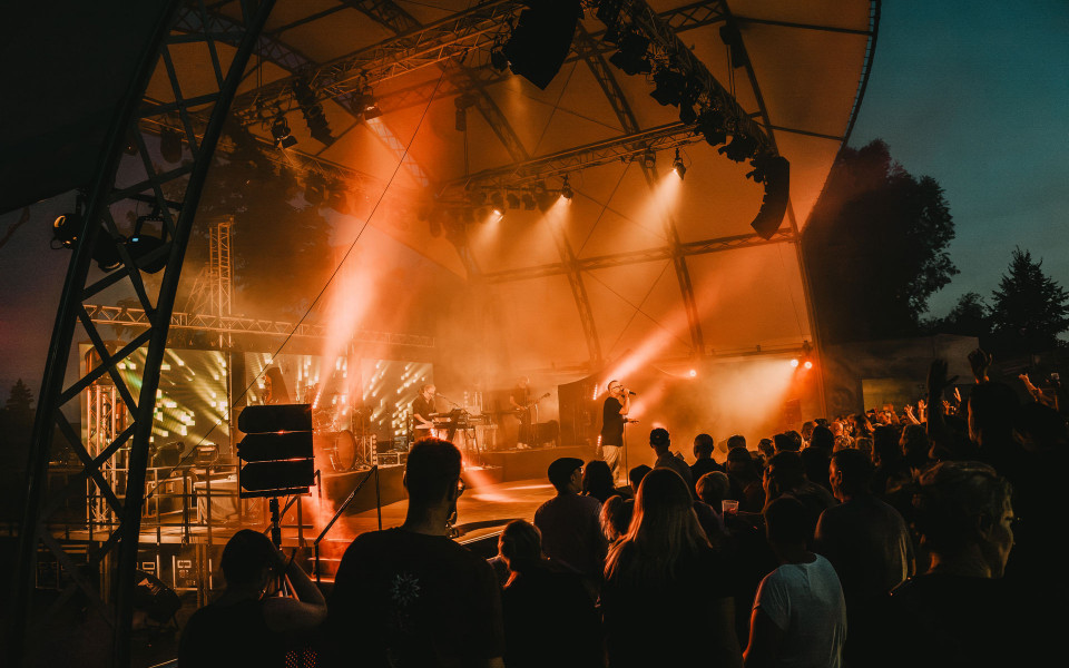 Auf einer Open-Air-Bühne spielt eine Band vor einem dicht gedrängten Publikum. Orange Scheinwerfer und Nebel tauchen die Szene bei Dämmerung in eine stimmungsvolle Konzertatmosphäre.