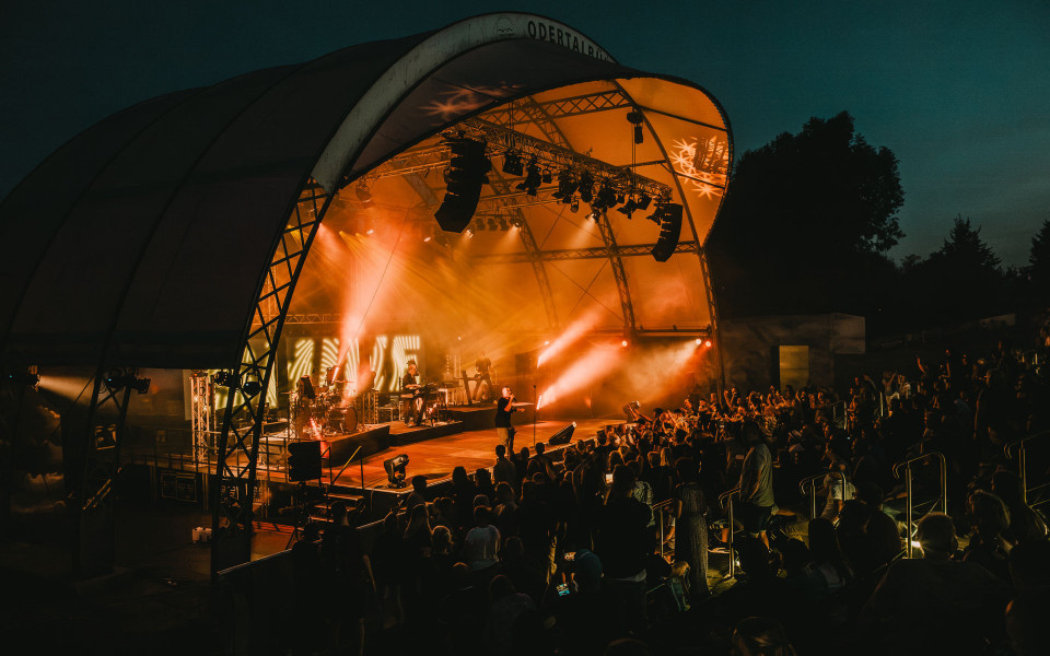 Auf einer überdachten Freilichtbühne findet bei Dunkelheit ein Konzert mit intensiven orangefarbenen Lichteffekten und Nebel statt. Vor der Bühne steht und sitzt ein großes Publikum, das den Auftritt der Band begeistert verfolgt.