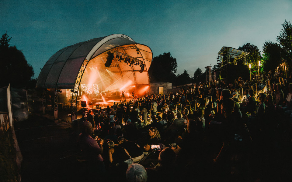 Bei Einbruch der Dunkelheit findet in einem Freilufttheater ein Konzert mit hell erleuchteter Bühne und orangefarbenem Scheinwerferlicht statt. Zahlreiche Zuschauer sitzen und stehen auf den Rängen und verfolgen die Aufführung unter dem offenen Himmel.