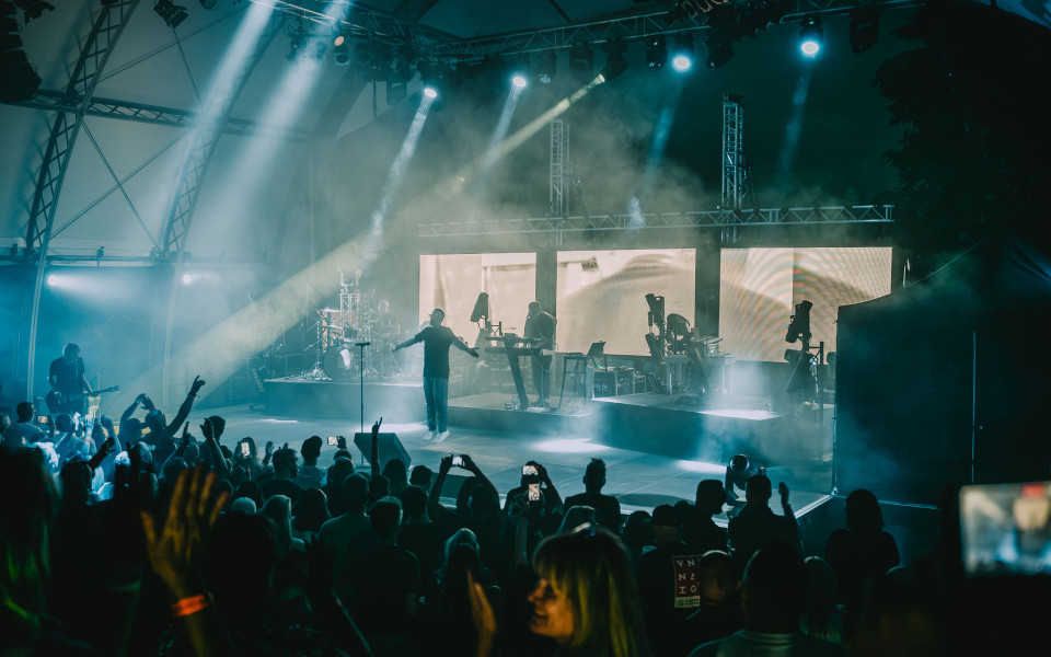 Auf einer großen Konzertbühne steht eine Band im Nebel, während blaue und weiße Scheinwerfer über das Publikum strahlen. Viele Zuschauer halten die Hände und Smartphones hoch und verfolgen die energiegeladene Live-Show.