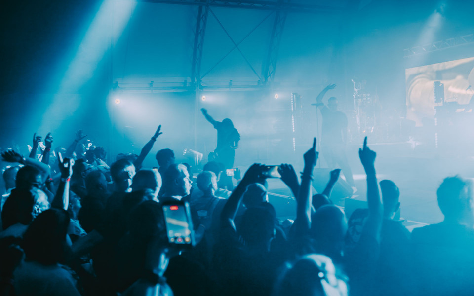 Ein Konzertsaal ist in blaues Licht und Nebel getaucht, während die Menge vor der Bühne die Arme hebt. Auf der Bühne sind die Silhouetten von Musikern und Instrumenten zu erkennen, und mehrere Zuschauer filmen mit ihren Smartphones.