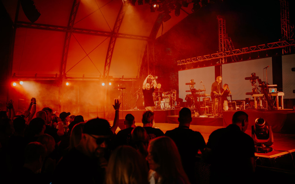 Auf einer Konzertbühne spielt eine Band unter intensiv rotem und orangefarbenem Licht, während Nebel die Szene durchzieht. Vor der Bühne steht ein dichtes Publikum, einige Menschen heben die Hände und feiern zur Musik.