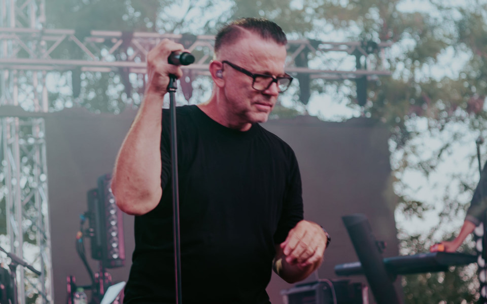 Ein Mann mit Brille und schwarzem Shirt steht auf einer Bühne und hält ein Mikrofon am Ständer. Im Hintergrund sind Bühnenbeleuchtung, Technik und unscharfe Bäume zu sehen.