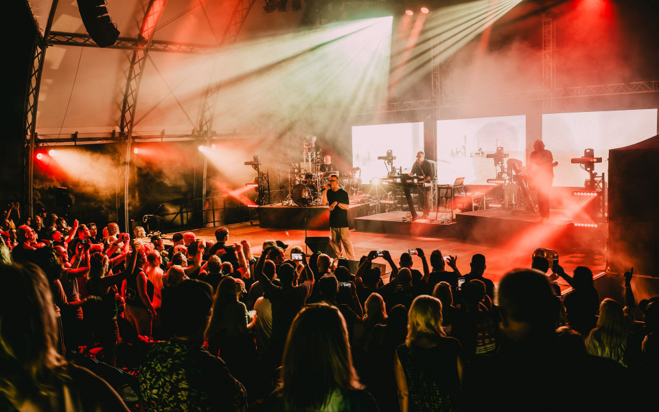 Auf einer großen Konzertbühne spielt eine Band vor einem dicht gedrängten Publikum, während viele Zuschauer ihre Handys hochhalten. Rote und weiße Scheinwerfer sowie Nebel erzeugen eine dramatische, energiegeladene Atmosphäre.