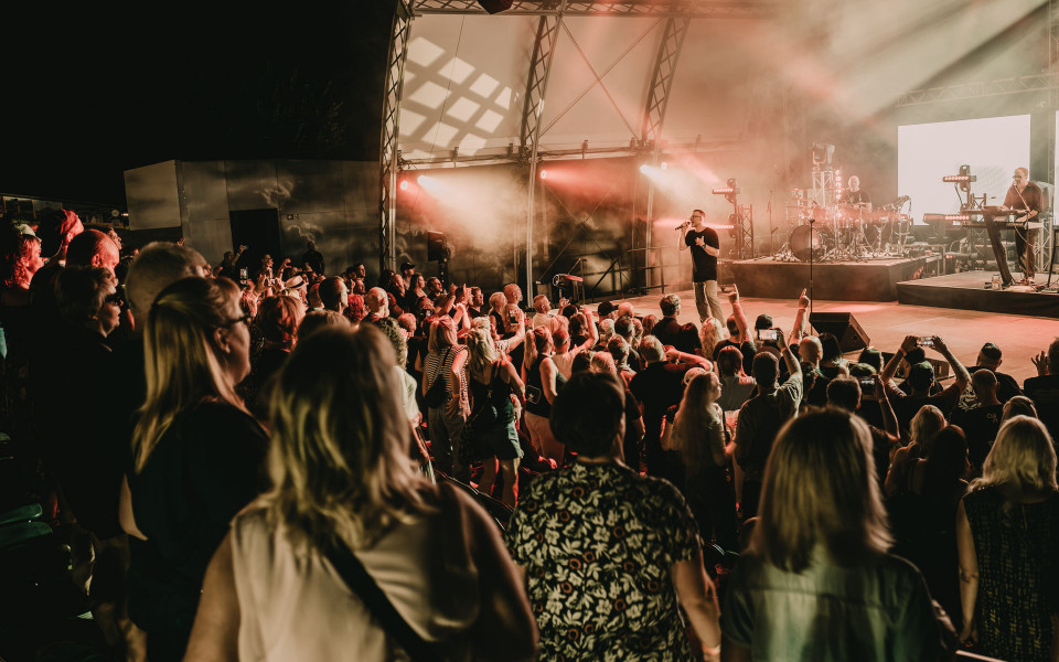 Eine große Menschenmenge steht bei einem Open-Air-Konzert vor einer hell beleuchteten Bühne. Der Sänger tritt im Schein von roten und weißen Scheinwerfern auf, während Musiker im Hintergrund spielen.