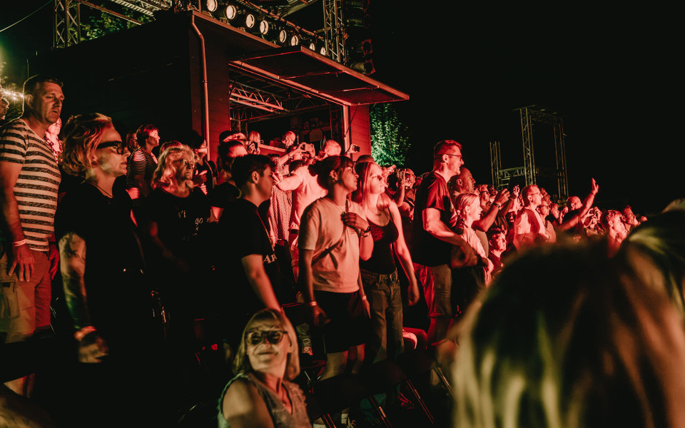 Eine Menschenmenge steht nachts bei einem Konzert vor einer beleuchteten Bühne und wird von rotem Licht angestrahlt. Viele Zuschauer schauen nach vorne, einige halten Handys hoch und wirken aufmerksam oder begeistert.