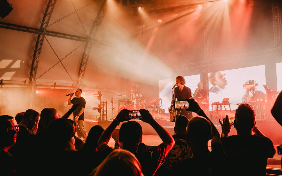 Auf einer Bühne performt eine Band vor einem dichten Publikum, während rote und orangefarbene Scheinwerfer durch den Nebel strahlen. Mehrere Zuschauer halten Smartphones hoch und filmen den Auftritt.