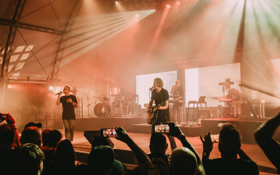 Eine Band spielt auf einer großen Bühne, die von warmen orangefarbenen Scheinwerfern und dichtem Nebel beleuchtet wird. Im Vordergrund steht das Publikum, einige halten Smartphones hoch und filmen den Auftritt.