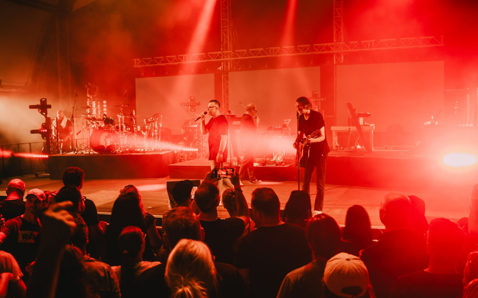 Eine Band tritt auf einer Bühne vor Publikum auf, die in intensives rotes Licht und Nebel getaucht ist. Im Vordergrund stehen Zuschauer, während Sänger, Gitarrist und Schlagzeuger unter Scheinwerfern performen.