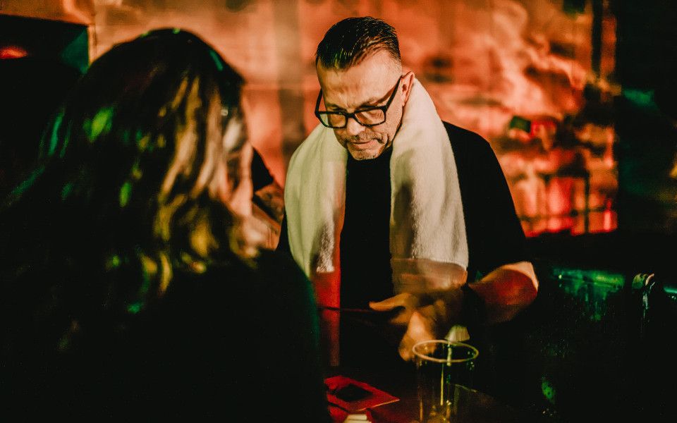 Ein Mann mit Brille und hellem Schal sitzt in einer dunkel beleuchteten Bar an einem Tisch. Im Vordergrund sind unscharf weitere Personen und Gläser zu sehen, während der Hintergrund warm orange und grün beleuchtet ist.
