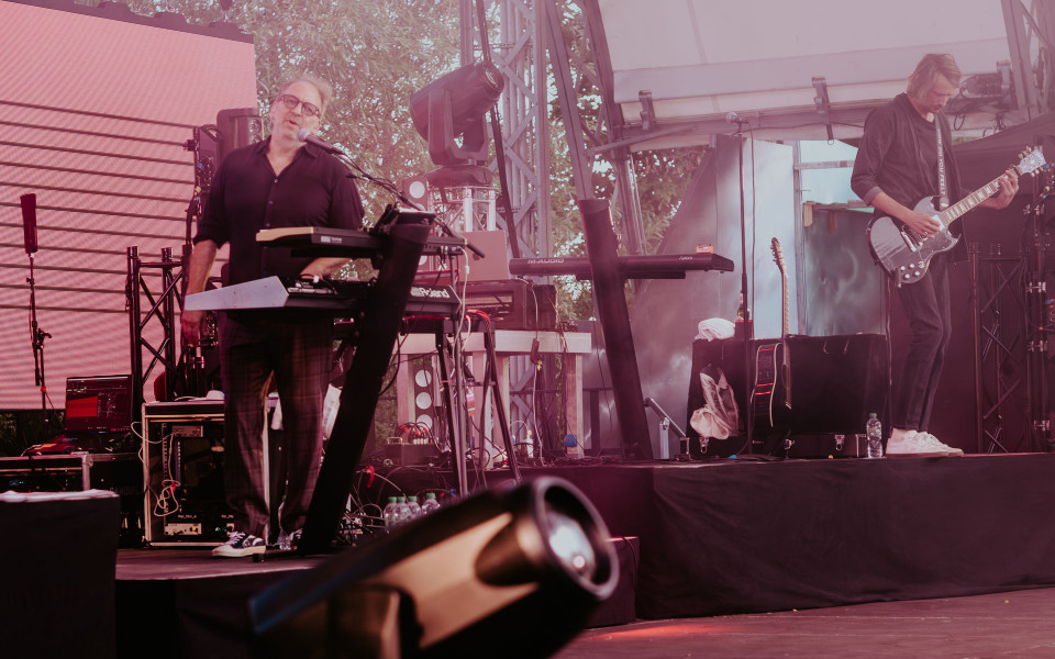 Zwei Musiker treten auf einer Open-Air-Bühne auf, einer singt und spielt Keyboard, während der andere E-Gitarre spielt. Die Szene ist in rosa Bühnenlicht getaucht, mit Technik, Mikrofonen und Instrumenten im Hintergrund.
