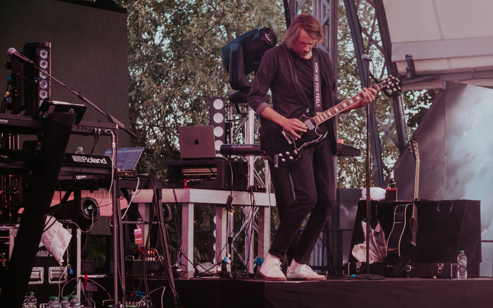 Ein Musiker spielt auf einer Open-Air-Bühne eine schwarze E-Gitarre, umgeben von Mikrofonen, Keyboards und Veranstaltungstechnik. Im Hintergrund sind Bäume, Scheinwerfer und weiteres Equipment zu sehen, während die Szene in rötlichem Bühnenlicht gehalten ist.