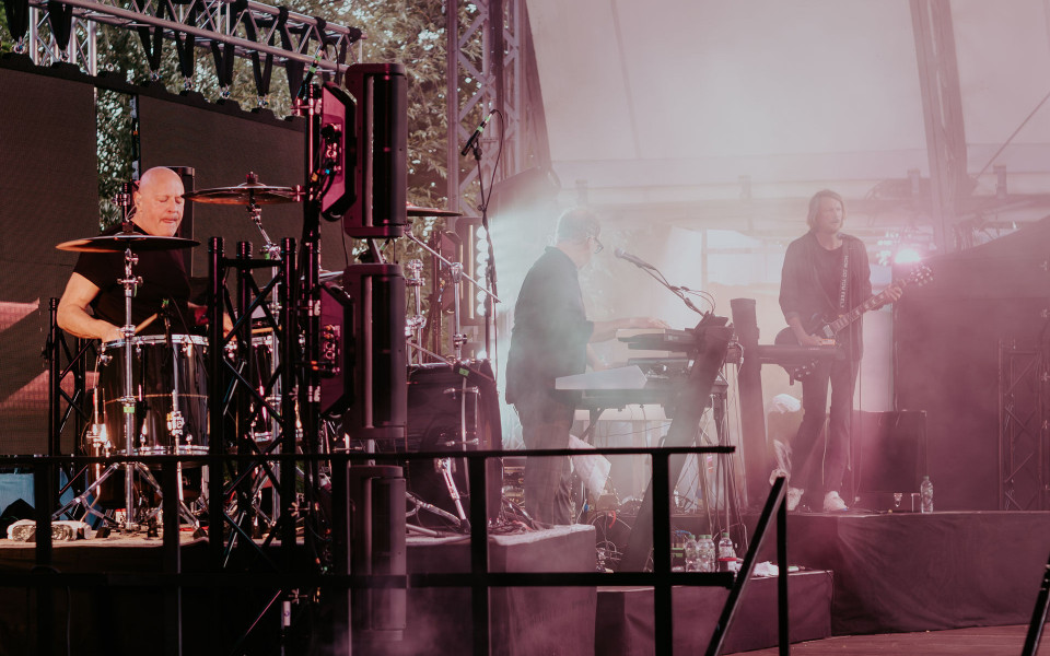 Eine Band spielt auf einer Bühne mit Schlagzeug, Keyboard und Gitarre, umgeben von Scheinwerfern und Bühnentechnik. Rosa-weißer Nebel und Lichtstrahlen erzeugen eine stimmungsvolle Konzertatmosphäre.