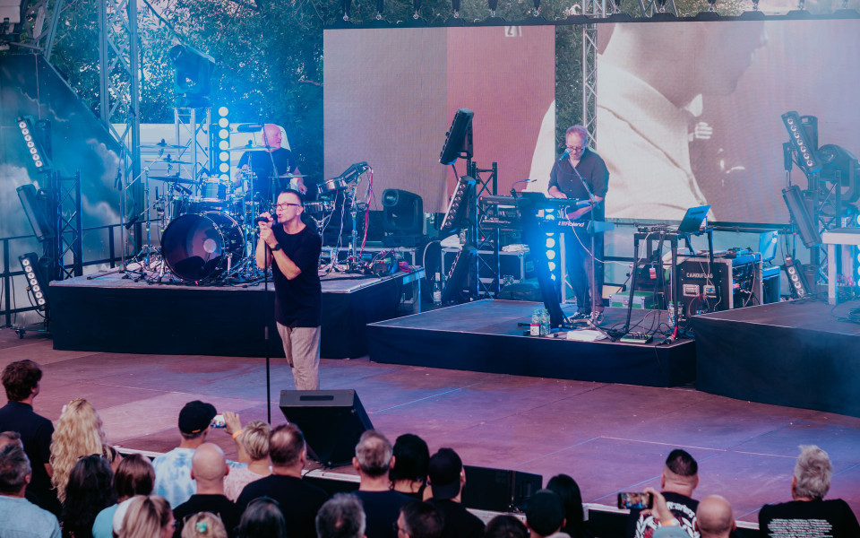 Eine Band tritt auf einer Freiluftbühne vor Publikum auf, mit Sänger am Mikrofon, Schlagzeuger und Keyboarder. Blaue Bühnenlichter, große LED-Leinwände und technische Ausrüstung prägen die Konzertatmosphäre.
