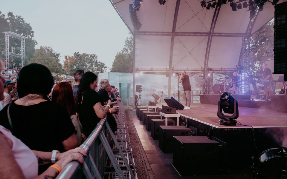Ein Sänger steht auf einer großen Open-Air-Bühne und singt vor einem Publikum, während farbiges Licht und Nebel die Szene prägen. Links drängen sich Zuschauer an der Absperrung, rechts sind Bandmitglieder, Instrumente und Lautsprecher unter dem Bühnendach zu sehen.