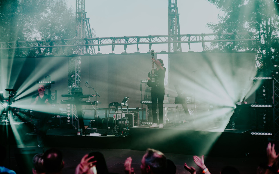 Auf einer Open-Air-Bühne spielt ein Musiker Gitarre, während helle Scheinwerferstrahlen durch Nebel über die Bühne schneiden. Im Vordergrund applaudiert das Publikum, und im Hintergrund sind weitere Instrumente, Lichttechnik und Bäume zu sehen.