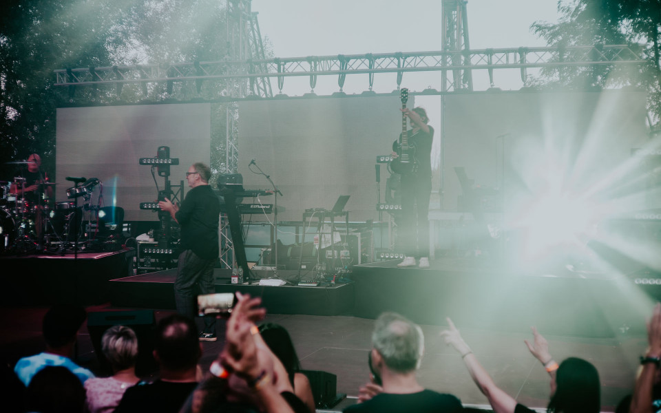 Auf einer Freiluftbühne spielt eine Band vor einem Publikum, während helle Scheinwerfer und Nebel die Szene durchziehen. Ein Gitarrist hält sein Instrument in die Höhe, links ist ein Schlagzeuger zu sehen und im Vordergrund klatschen Zuschauer.