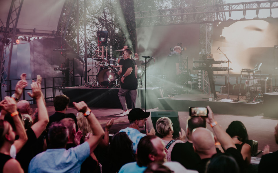Auf einer Open-Air-Bühne spielt eine Band vor einem begeisterten Publikum, das die Hände hebt und klatscht. Bunte Bühnenlichter, Nebel und Musikinstrumente wie Schlagzeug und Keyboard prägen die lebhafte Konzertatmosphäre.