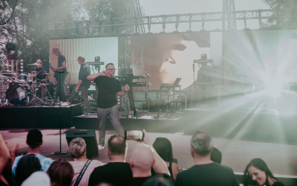Auf einer Open-Air-Bühne treten Musiker vor einem Publikum auf, während helle Scheinwerfer und Nebel die Szene durchziehen. Im Hintergrund sind große LED-Leinwände, Instrumente und Bühnentechnik zu sehen.