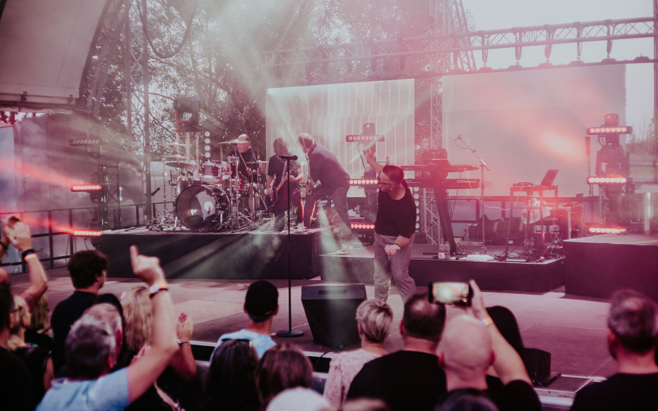Eine Band tritt auf einer Open-Air-Bühne mit Schlagzeug, Gitarren und Keyboard auf, während rosa Licht und Nebel die Szene füllen. Vor der Bühne steht ein Publikum, das jubelt, die Arme hebt und mit Handys filmt.