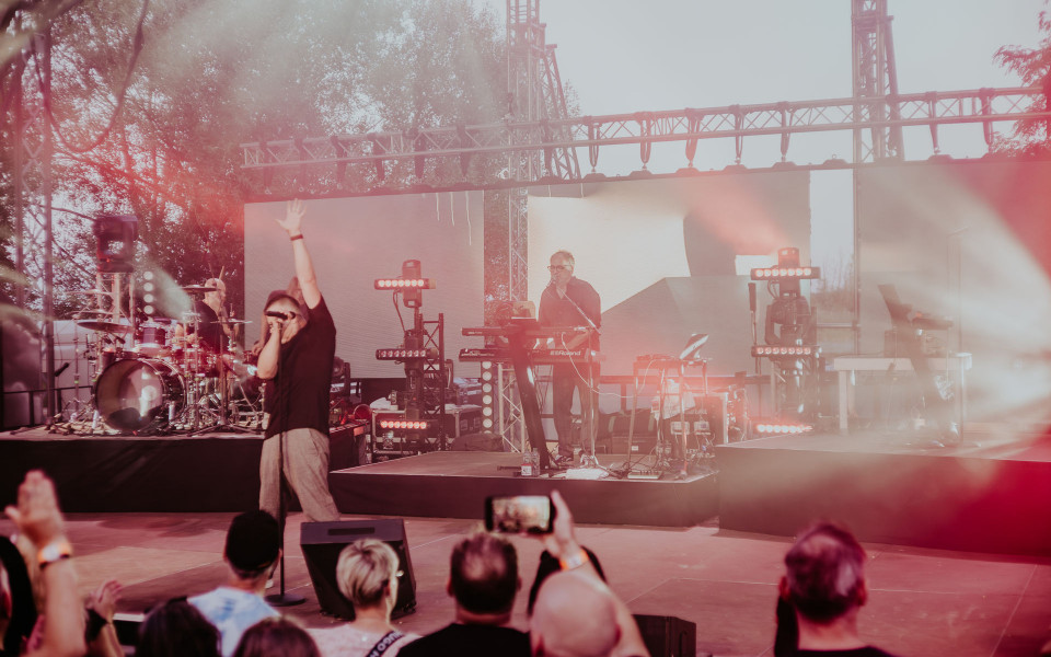 Eine Band tritt auf einer Open-Air-Bühne vor Publikum auf, während der Sänger mit erhobenem Arm ins Mikrofon singt. Rotes Bühnenlicht, Nebel und große LED-Wände sorgen für eine intensive Konzertatmosphäre.