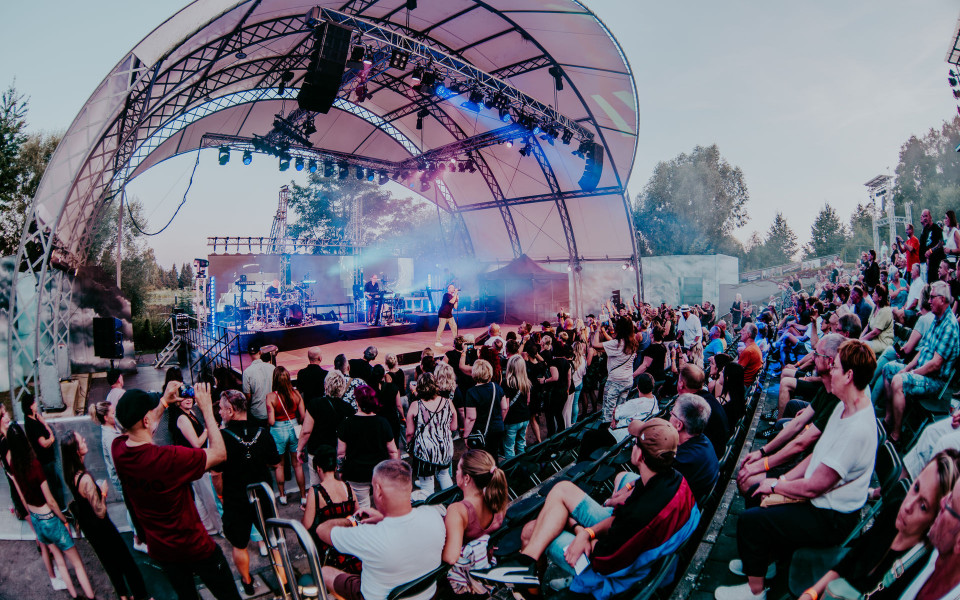 Eine Band tritt auf einer großen überdachten Freilichtbühne mit bunten Scheinwerfern und Bühnennebel auf. Das Publikum sitzt und steht dicht gedrängt in einem Amphitheater und verfolgt das Konzert bei Tageslicht.