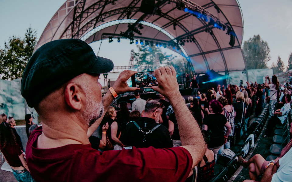 Ein Mann mit schwarzer Mütze filmt mit seinem Smartphone ein Open-Air-Konzert vor einer großen überdachten Bühne. Um ihn herum stehen und sitzen viele Zuschauer, während blaues Bühnenlicht und die Abendstimmung die Szene prägen.