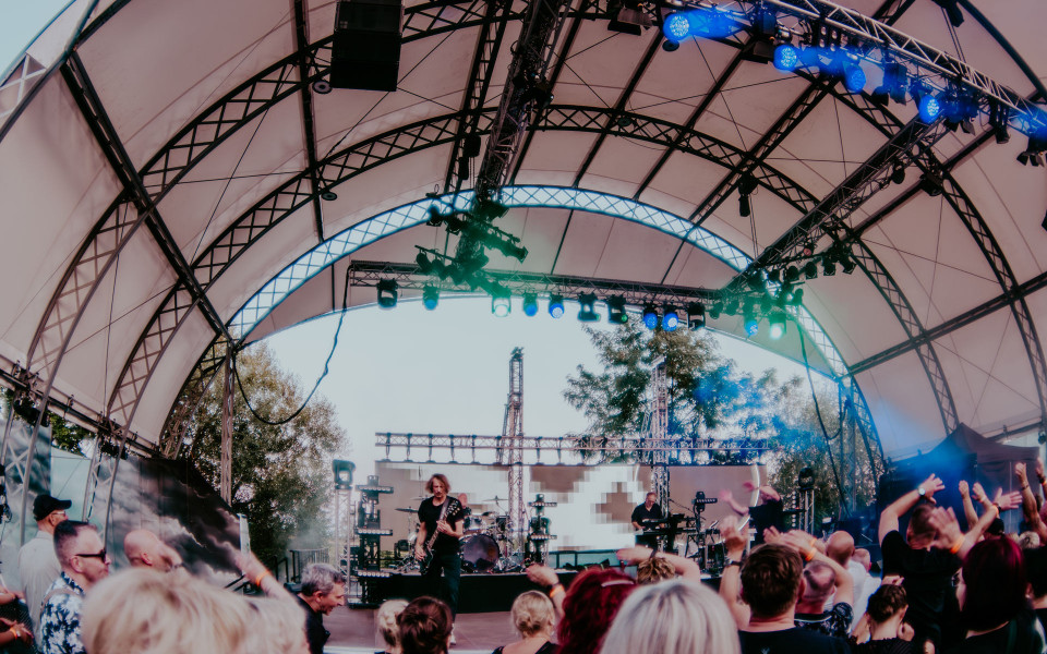 Eine Band spielt auf einer überdachten Open-Air-Bühne mit vielen Scheinwerfern und blauem Licht. Vor der Bühne steht ein Publikum mit erhobenen Händen und feiert das Konzert.