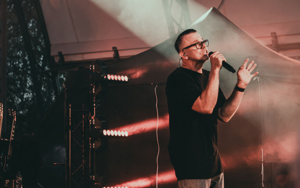 Ein Mann mit Brille singt auf einer Bühne in ein Mikrofon, während rotes und weißes Licht durch Nebel strahlt. Im Hintergrund sind Bühnentechnik und Instrumente zu sehen, was eine Konzertatmosphäre erzeugt.
