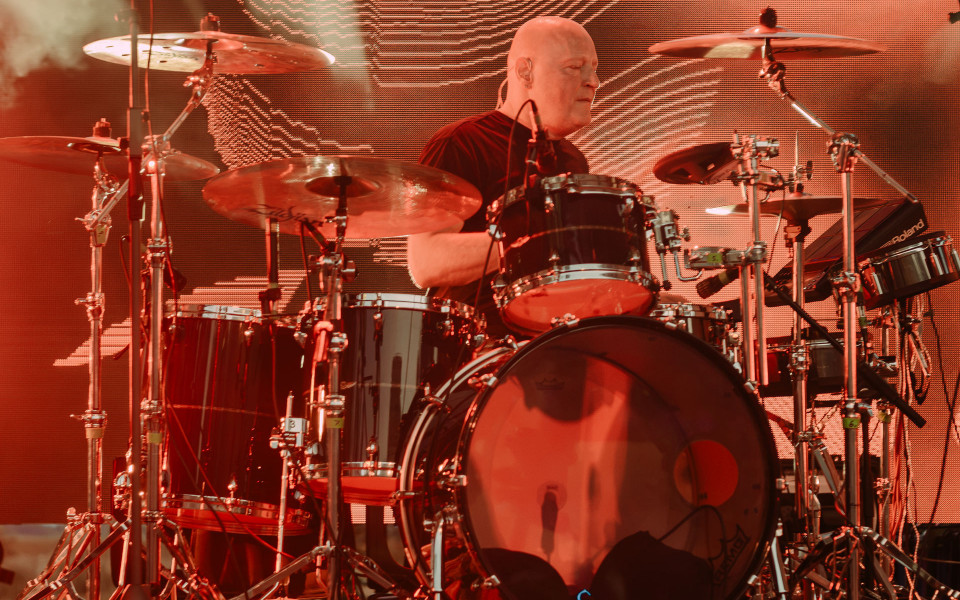 Ein Schlagzeuger sitzt auf einer Bühne hinter einem großen Drumset und spielt konzentriert unter rotem Licht. Im Hintergrund leuchtet eine große LED-Wand, während Becken, Trommeln und Mikrofone die Szene dominieren.