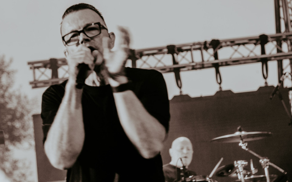 Ein Sänger mit Brille steht unscharf im Vordergrund auf einer Bühne und hält ein Mikrofon in der Hand. Im Hintergrund sitzt ein Schlagzeuger an seinem Drumset, während Bühnenbeleuchtung und Metalltraversen zu sehen sind.