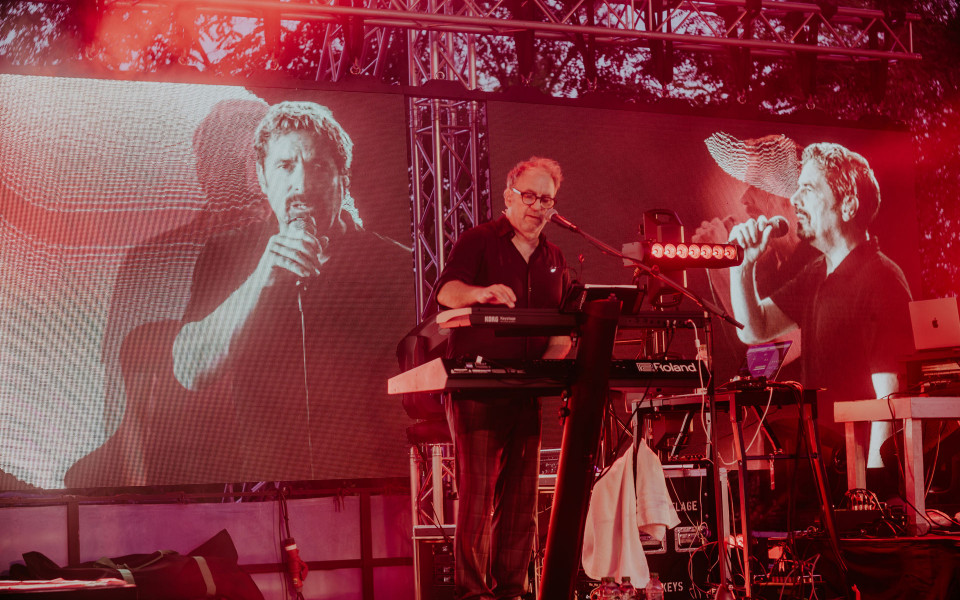 Ein Musiker steht auf einer rot beleuchteten Bühne und spielt Keyboard vor einem Mikrofon. Hinter ihm zeigen große LED-Leinwände schwarz-weiße Aufnahmen von singenden Personen.