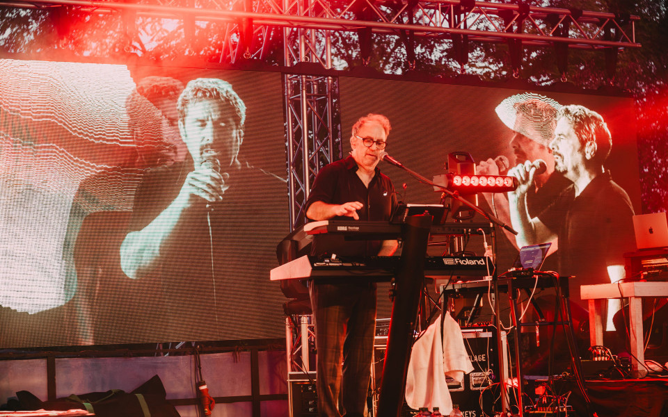 Ein Musiker steht auf einer rot beleuchteten Bühne und singt an einem Mikrofon, während er vor mehreren Keyboards steht. Hinter ihm zeigen große LED-Leinwände Nahaufnahmen von singenden Personen, was die Konzertatmosphäre verstärkt.