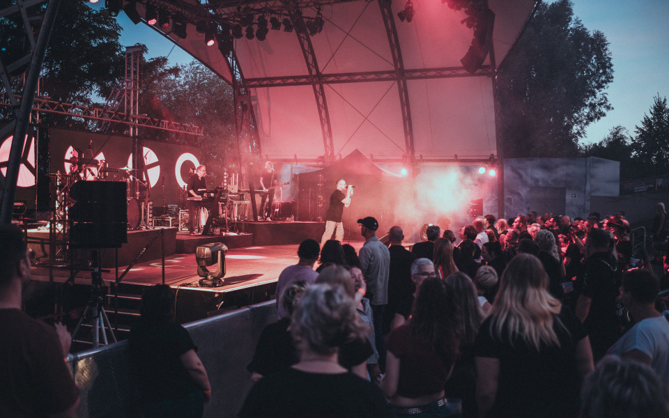 Eine Band tritt auf einer überdachten Open-Air-Bühne vor einem dicht gedrängten Publikum auf. Rotes Bühnenlicht und Nebel schaffen eine stimmungsvolle Konzertatmosphäre in der Abenddämmerung.