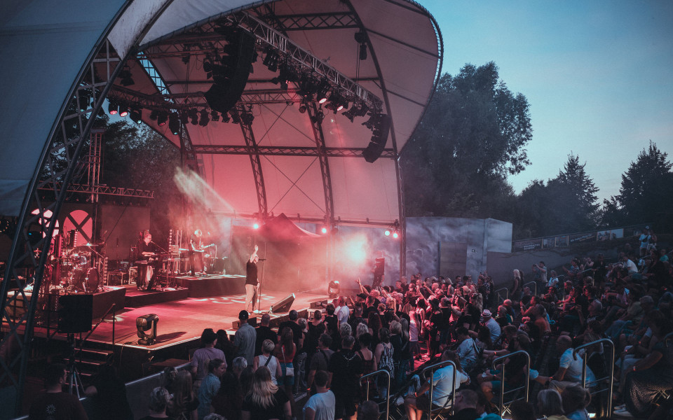 Auf einer überdachten Freilichtbühne spielt eine Band vor einem großen Publikum, während rotes Licht und Nebel die Szene dramatisch beleuchten. Die Zuschauer stehen und sitzen dicht gedrängt vor der Bühne, umgeben von Bäumen in der Abenddämmerung.