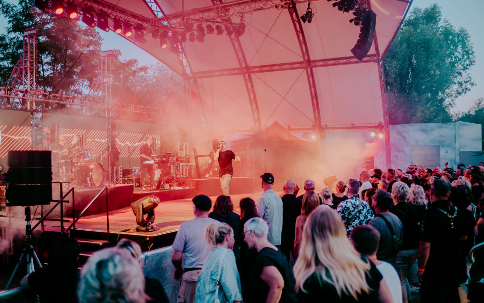 Auf einer überdachten Open-Air-Bühne tritt eine Band vor einem dicht gedrängten Publikum auf. Rotes und blaues Licht sowie Nebel tauchen die Szene in eine lebendige Konzertatmosphäre.