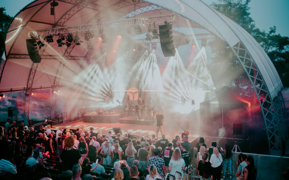Eine große Open-Air-Bühne ist in dichten Nebel und helle Lichtstrahlen gehüllt, während Musiker auftreten. Vor der Bühne steht und sitzt ein großes Publikum, das die Konzertatmosphäre genießt.