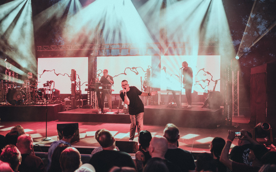 Ein Sänger steht mit Mikrofon im Vordergrund einer hell beleuchteten Konzertbühne, während die Band hinter ihm spielt. Vor der Bühne schaut ein Publikum zu, umgeben von starken Scheinwerfern, Nebel und großen LED-Wänden.