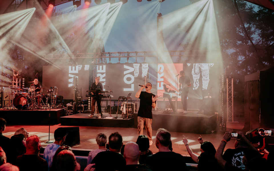 Eine Band spielt auf einer hell erleuchteten Bühne mit starkem Scheinwerferlicht und Nebel, während der Sänger vorne ins Mikrofon singt. Vor der Bühne steht ein Publikum, das dem Konzert zuschaut und teilweise die Hände hebt oder mit dem Handy filmt.