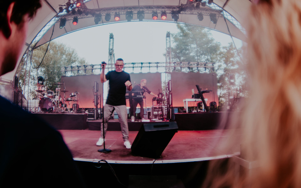 Auf einer überdachten Open-Air-Bühne steht ein Sänger mit Mikrofon vor Instrumenten, Lichtanlagen und großen LED-Wänden. Im Vordergrund sind unscharf zwei Zuschauer zu sehen, wodurch der Eindruck entsteht, direkt aus dem Publikum auf die Bühne zu blicken.