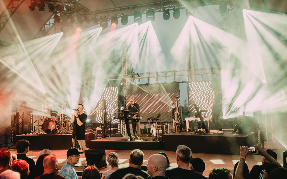 Eine Band spielt auf einer großen Open-Air-Bühne vor Publikum, während helle Scheinwerfer durch dichten Nebel strahlen. Im Hintergrund leuchten LED-Wände mit grafischen Mustern, und die Zuschauer verfolgen das Konzert dicht vor der Bühne.