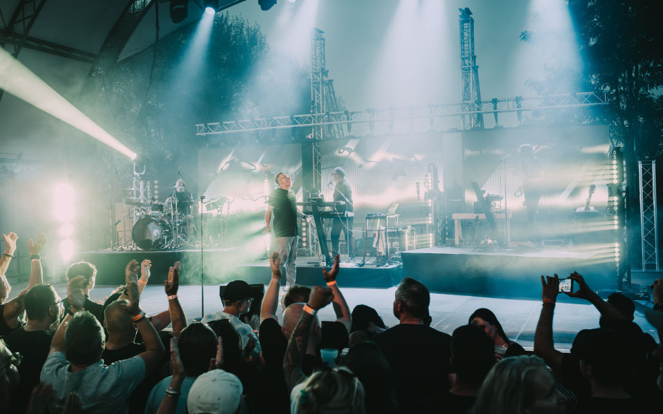 Auf einer Konzertbühne steht eine Band im dichten Nebel, während helle Scheinwerfer in Blau- und Weißtönen über die Bühne strahlen. Vor der Bühne jubelt ein Publikum mit erhobenen Händen, einige filmen den Auftritt mit ihren Smartphones.