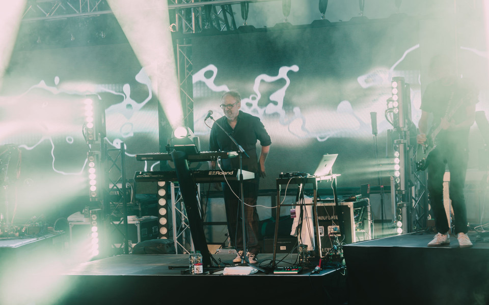Auf einer Konzertbühne steht ein Musiker an Keyboards und einem Mikrofon, während rechts ein Gitarrist im Nebel zu sehen ist. Helle Scheinwerfer und eine große LED-Wand mit abstrakten Mustern erzeugen eine atmosphärische, grünlich leuchtende Bühnenszene.