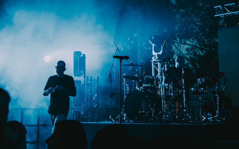 Auf einer blau beleuchteten Konzertbühne stehen Mikrofone, ein Schlagzeug und weitere Technik im dichten Nebel. Links bewegt sich ein Musiker im Vordergrund, während rechts ein Schlagzeuger im Hintergrund zu sehen ist.