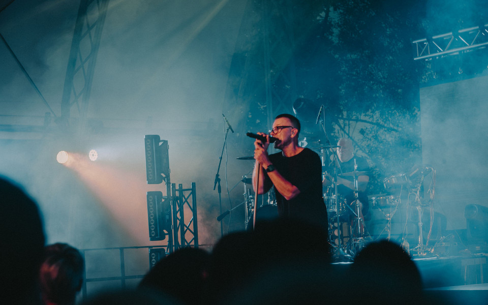 Ein Sänger steht bei einem Konzert auf einer nebligen Bühne und singt in ein Mikrofon. Im Hintergrund spielt ein Schlagzeuger, während blaue Lichter und die Silhouetten des Publikums die Atmosphäre prägen.