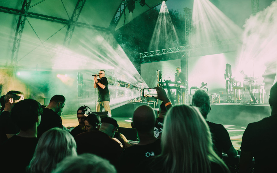 Ein Sänger steht auf einer nebligen Konzertbühne vor einem Publikum, während grüne und weiße Lichtstrahlen durch die Luft schneiden. Im Hintergrund spielt ein Musiker an Keyboards und elektronischem Equipment vor großen LED-Flächen und Bühnenträgern.