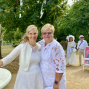Zwei Frauen in weißer Kleidung stehen lächelnd und eng umarmt in einem grünen Park. Im Hintergrund gehen weitere weiß gekleidete Personen an einem Plakat mit der Aufschrift „Love is in the air“ vorbei.