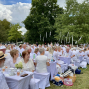 Viele Menschen sitzen bei einer festlichen Veranstaltung im Park an weiß gedeckten Tischen und tragen überwiegend weiße Kleidung. Zwischen den Bäumen hängen weiße Bänder, während unter einem bewölkten Himmel gemeinsam gegessen und getrunken wird.